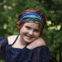 Girl with red hair and colorful headband smiling outdoors.