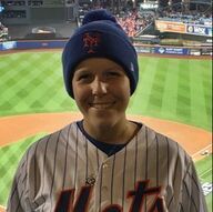 Person in Mets jersey and blue cap smiles at baseball stadium.