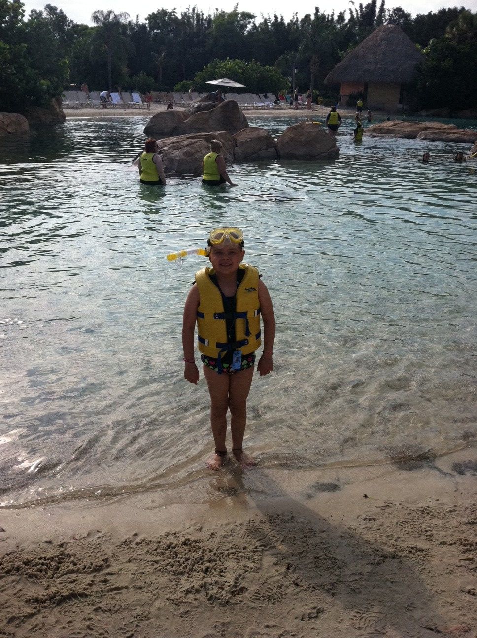Child in a yellow life vest and snorkel in shallow water at a beach, with rocks and huts in the background.