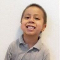 Boy with short hair smiling, wearing a gray polo shirt against a white wall.