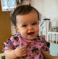 Smiling baby in a pink shirt with a star and teddy bear pattern in a hospital room.