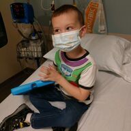 Child wearing a mask and Buzz Lightyear shirt, sitting on a hospital bed with a tablet.