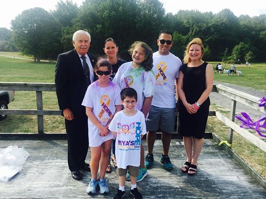 Group of people, some wearing tie-dye shirts, on a wooden platform outdoors. Some people have purple ribbons.