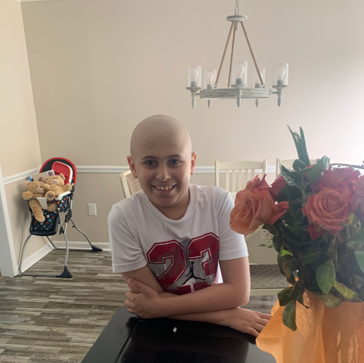 Smiling person with bald head wearing a white shirt, arms crossed, standing near a vase of roses; inside a home.