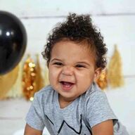 Smiling toddler with curly hair, wearing a gray shirt, in front of a black balloon and gold decorations.