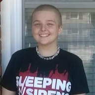Person with a shaved head smiles, wearing a black shirt and beaded necklace in front of a window.
