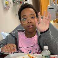 Boy in hospital bed raises hand; wears glasses, pink shirt, gray jacket; food tray nearby.