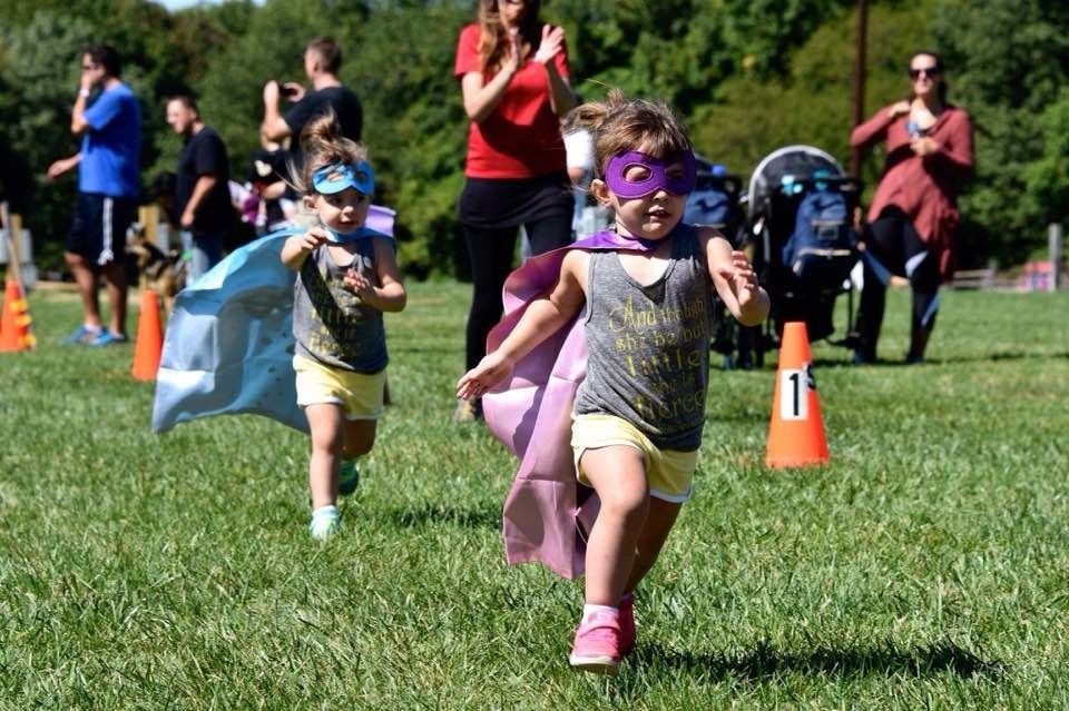 Two children in superhero capes and masks run on grass towards the camera. People watch in background.