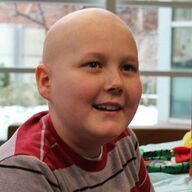 Smiling child with a shaved head wearing a red and white striped shirt, sitting indoors.