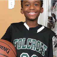Boy in a basketball jersey holding a ball, smiling. Jersey reads 