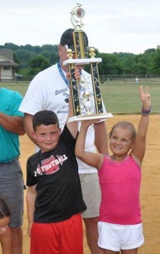 Two children holding trophy, celebrating a win. Boy in red shorts, girl in white, both smiling, outdoors.
