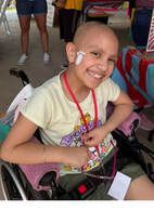 Smiling child in a wheelchair with medical equipment, wearing a yellow shirt and red lanyard, outside.