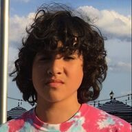 Person with curly dark hair and tie-dye shirt, looking directly at the camera outdoors with a blue sky background.