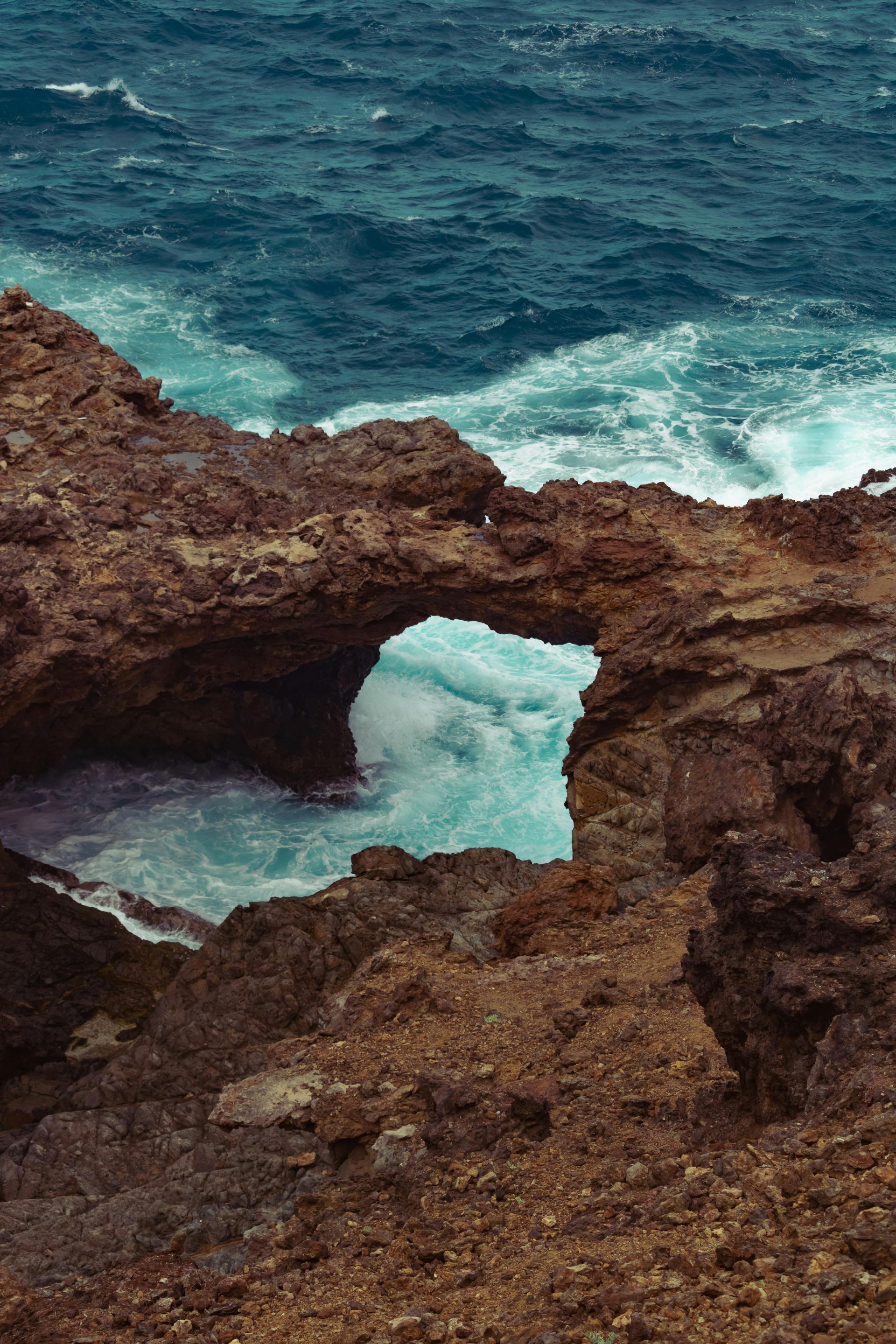 Sea arch carved in brown rocks with turquoise water rushing through.