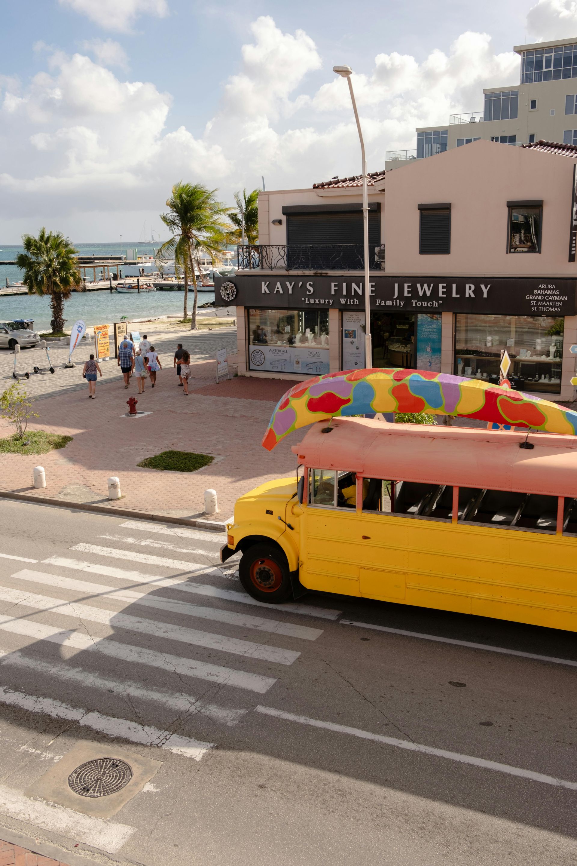 Yellow school bus with colorful canopy parked near jewelry store, sidewalk, and water.