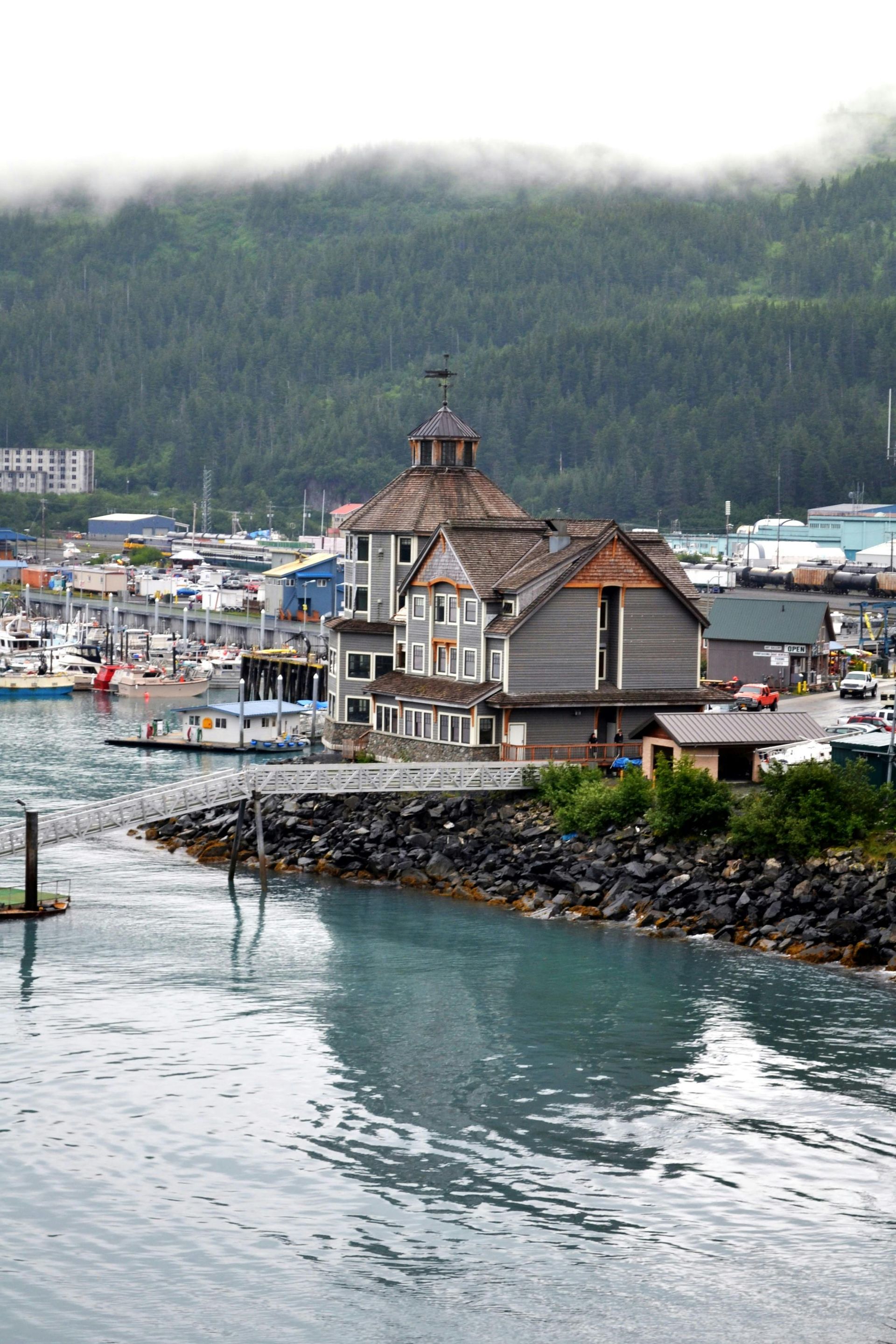 Wooden building on a rocky shoreline in a coastal town with boats and a forested hillside in the background.