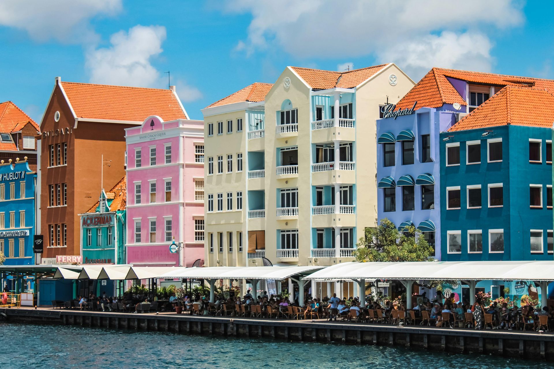 Colorful buildings line a waterfront. People sit under a white awning along the water.