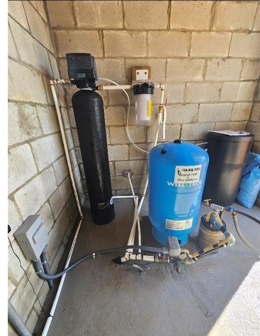 Well water filtration system with black filter tank, clear pre-filter, and blue pressure tank against a concrete wall. Well water filtration system with black filter tank, clear pre-filter, and blue pressure tank against a concrete wall.