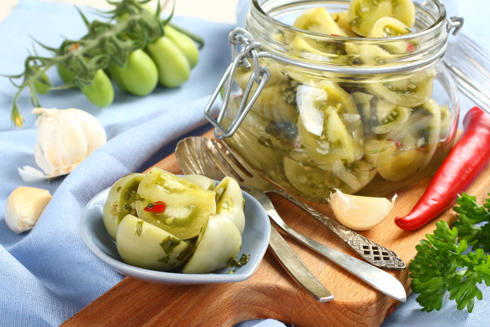 Pickled green tomatoes in a jar with a bunch of green tomatoes to the side.