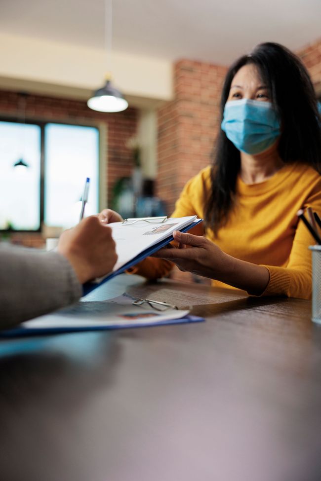 A woman wearing a mask is giving a clipboard to another woman.