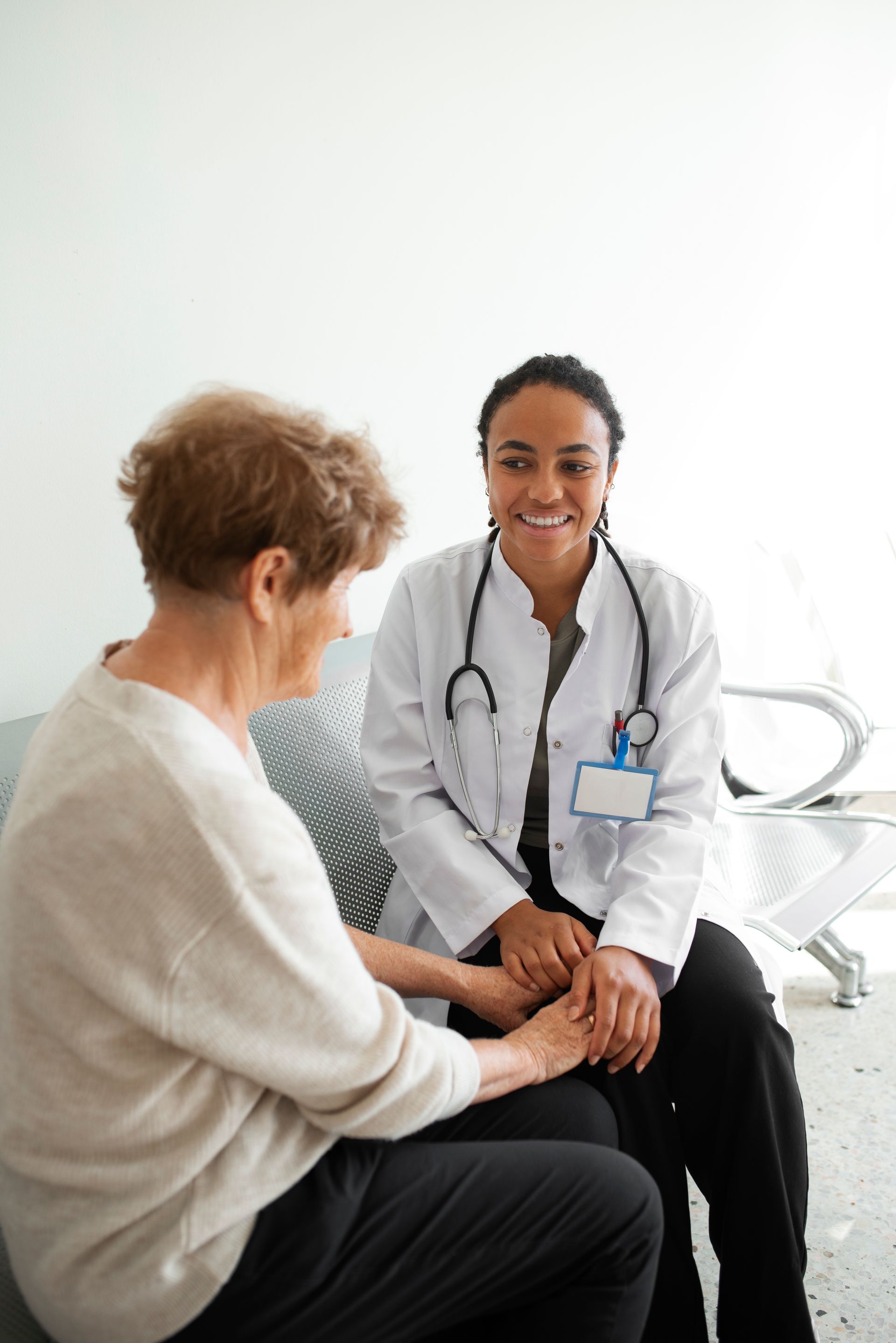 A doctor is holding the hand of an elderly woman while sitting on a couch.