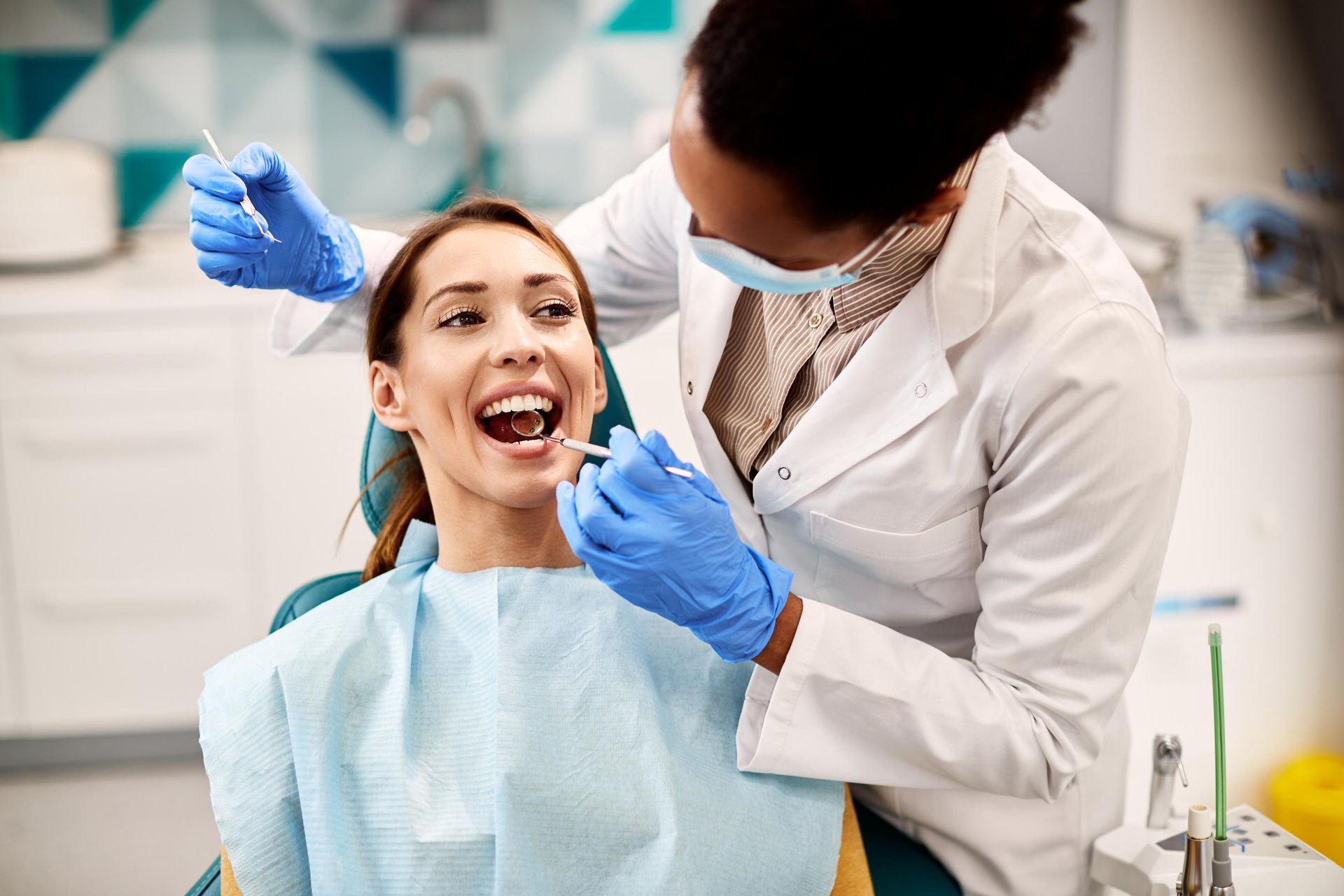 A dental professional in a white coat and blue gloves performs a check-up on a smiling patient in a dental chair.