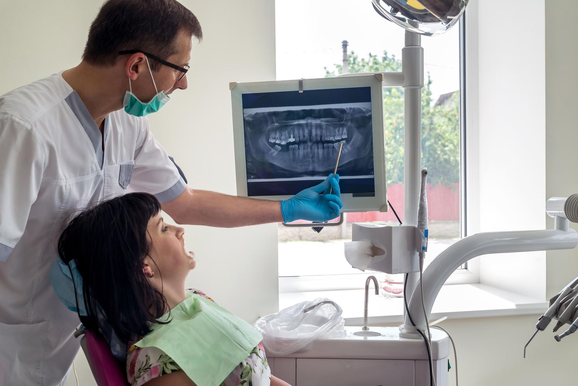 A dentist points to a digital panoramic X-ray on a monitor while explaining it to a patient sitting in a dental chair.