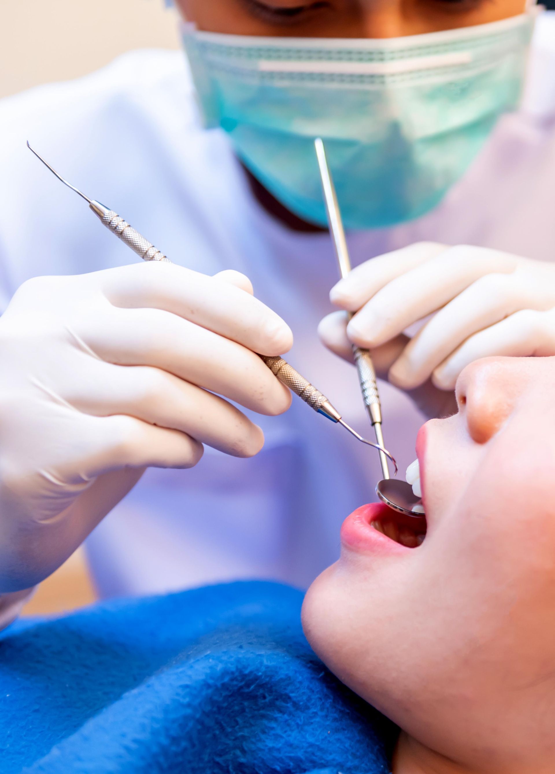 A dentist wearing a blue mask and white gloves uses a mirror and probe tool to examine a patient's open mouth.
