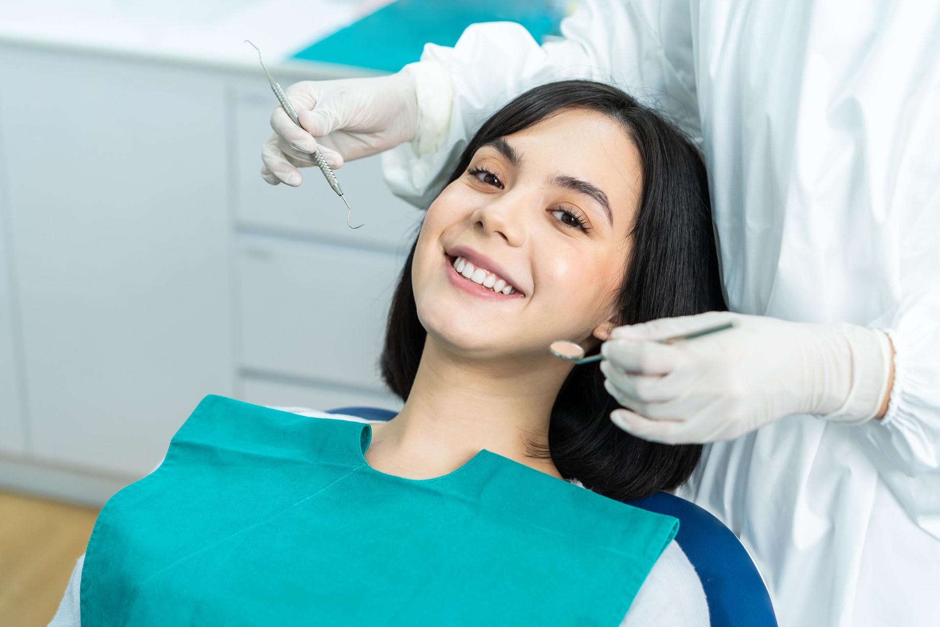 A smiling patient in a teal bib receiving a dental exam from a professional wearing white protective gear.