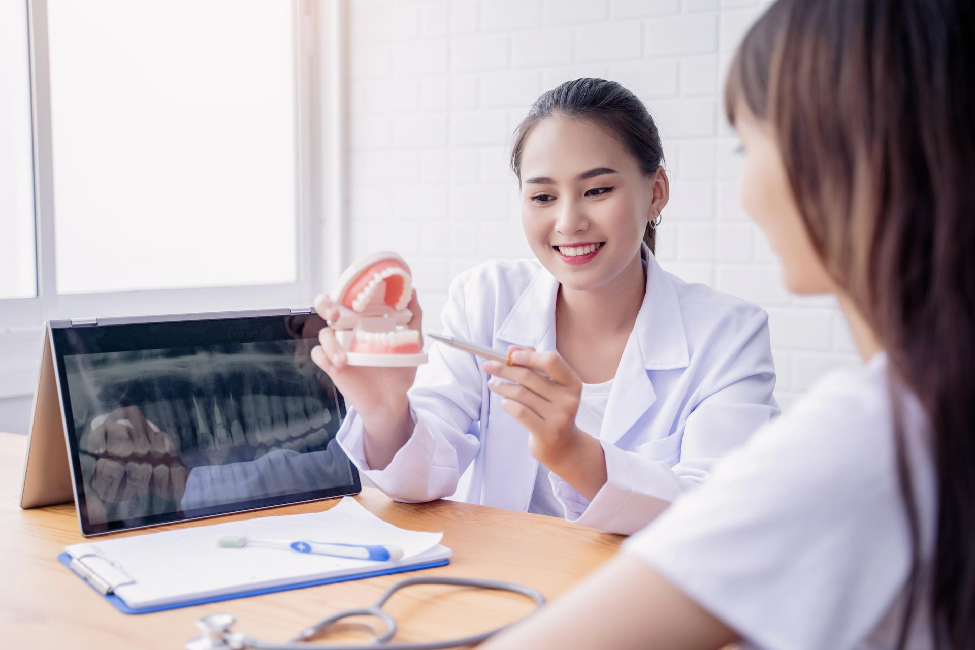 A dentist in a white coat smiles while explaining a dental model to a patient in an office with an X-ray on a laptop.