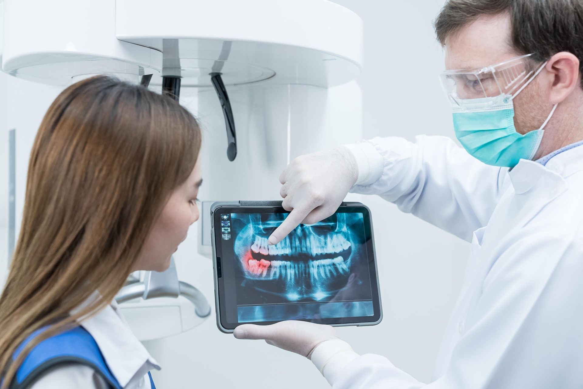 A dentist in a mask and glasses points to an x-ray on a tablet while discussing it with a patient in a dental office.