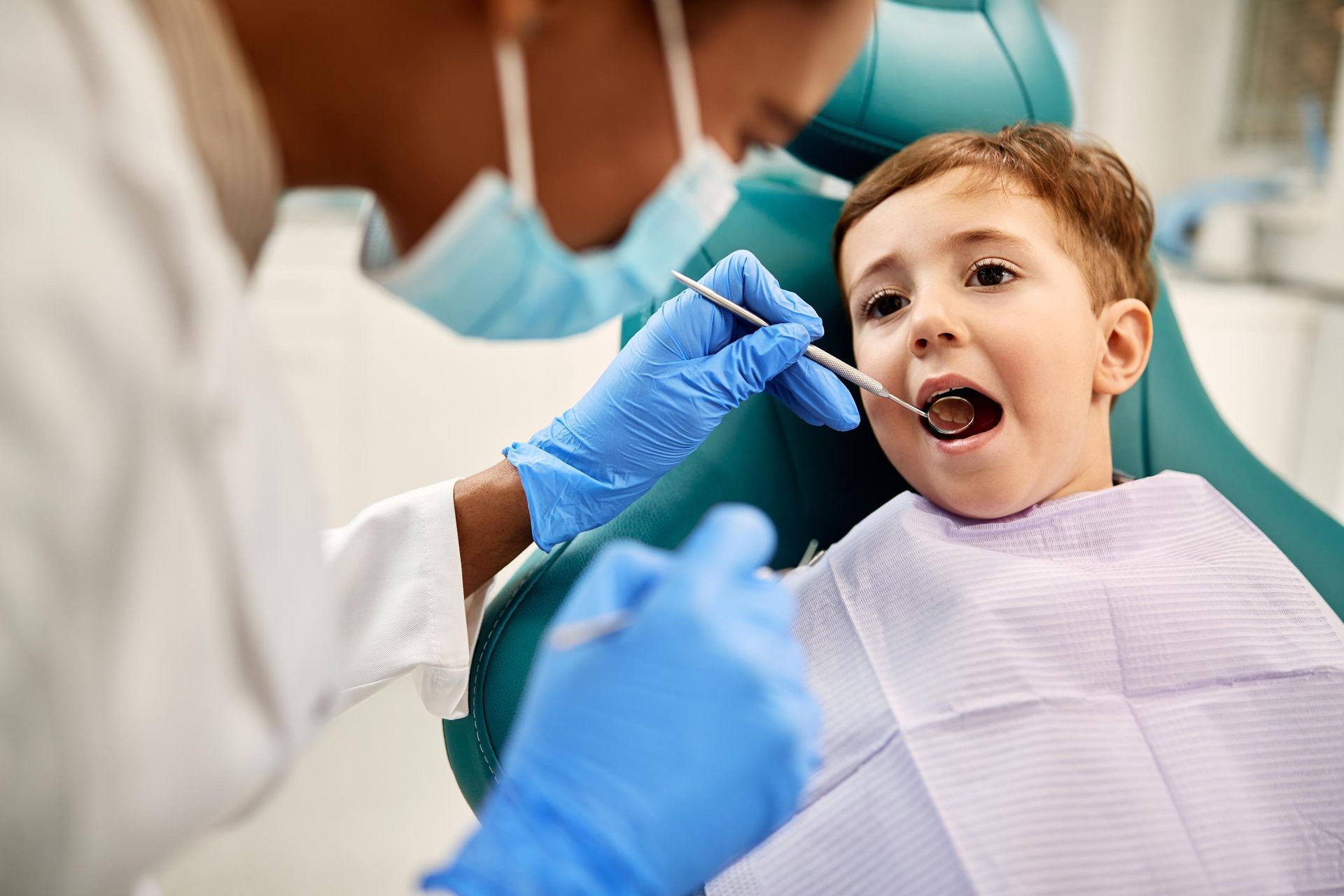 A dentist in scrubs and blue gloves examines a child’s teeth with a mouth mirror in a clinical setting.