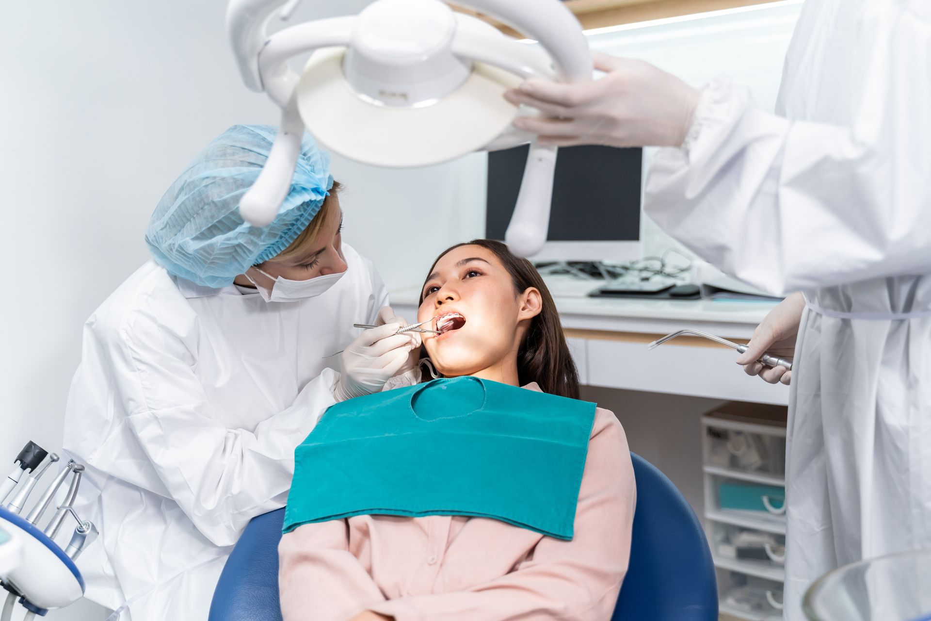 A dental professional examines a patient’s teeth while an assistant adjusts the overhead light in a clinical office.
