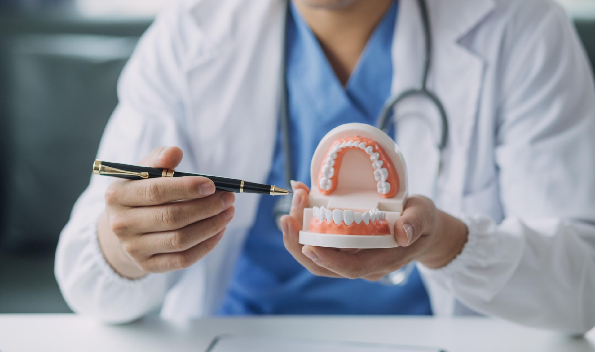 A dental professional in a white coat and blue scrubs holds a model of teeth and a pen, explaining a dental procedure.