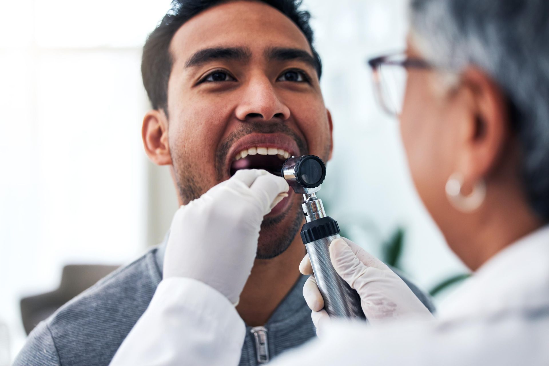 A healthcare professional in white gloves uses a lighted otoscope to examine a patient's mouth during a checkup.