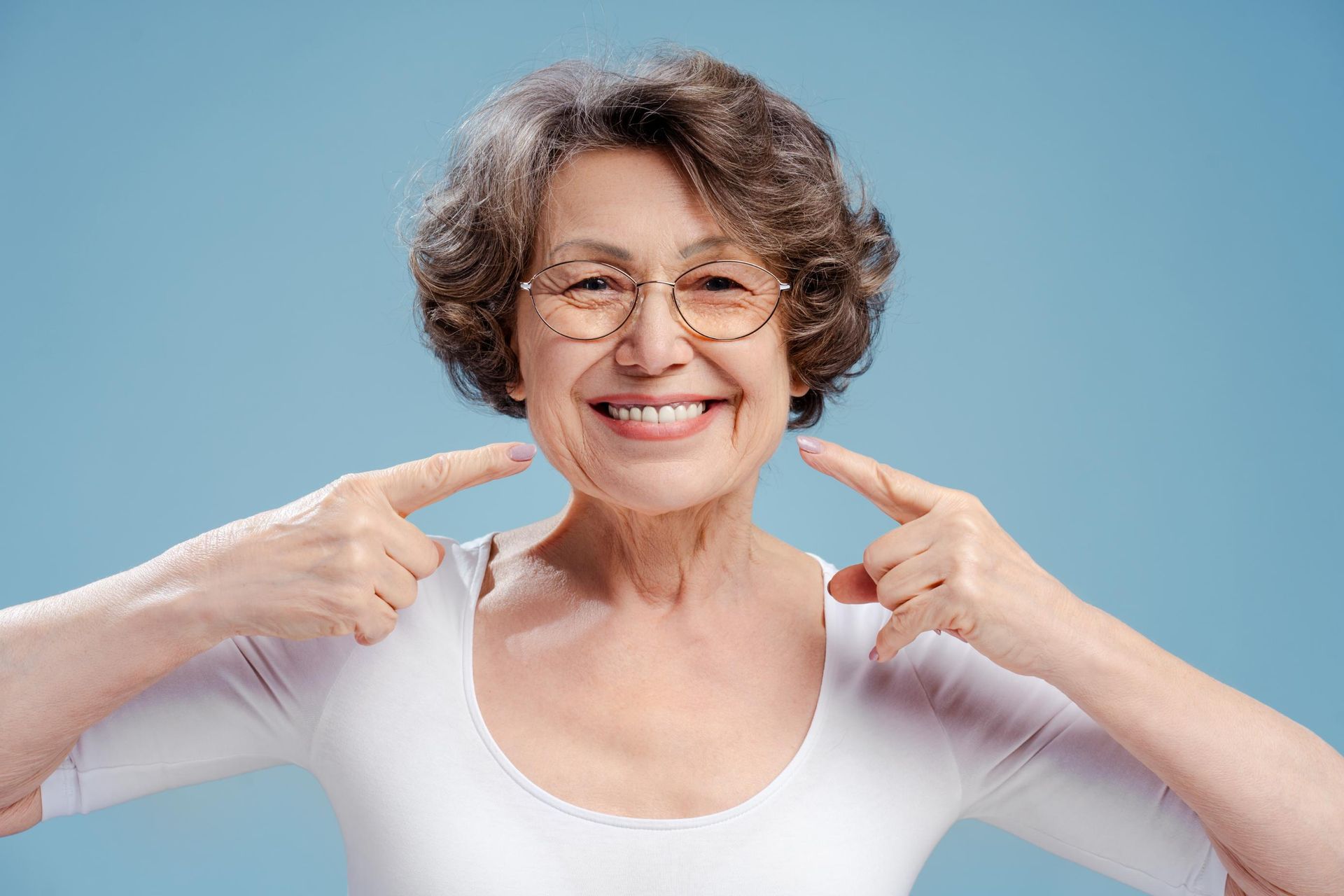 A person wearing glasses smiles at the camera, pointing with both index fingers toward their mouth against a blue background.