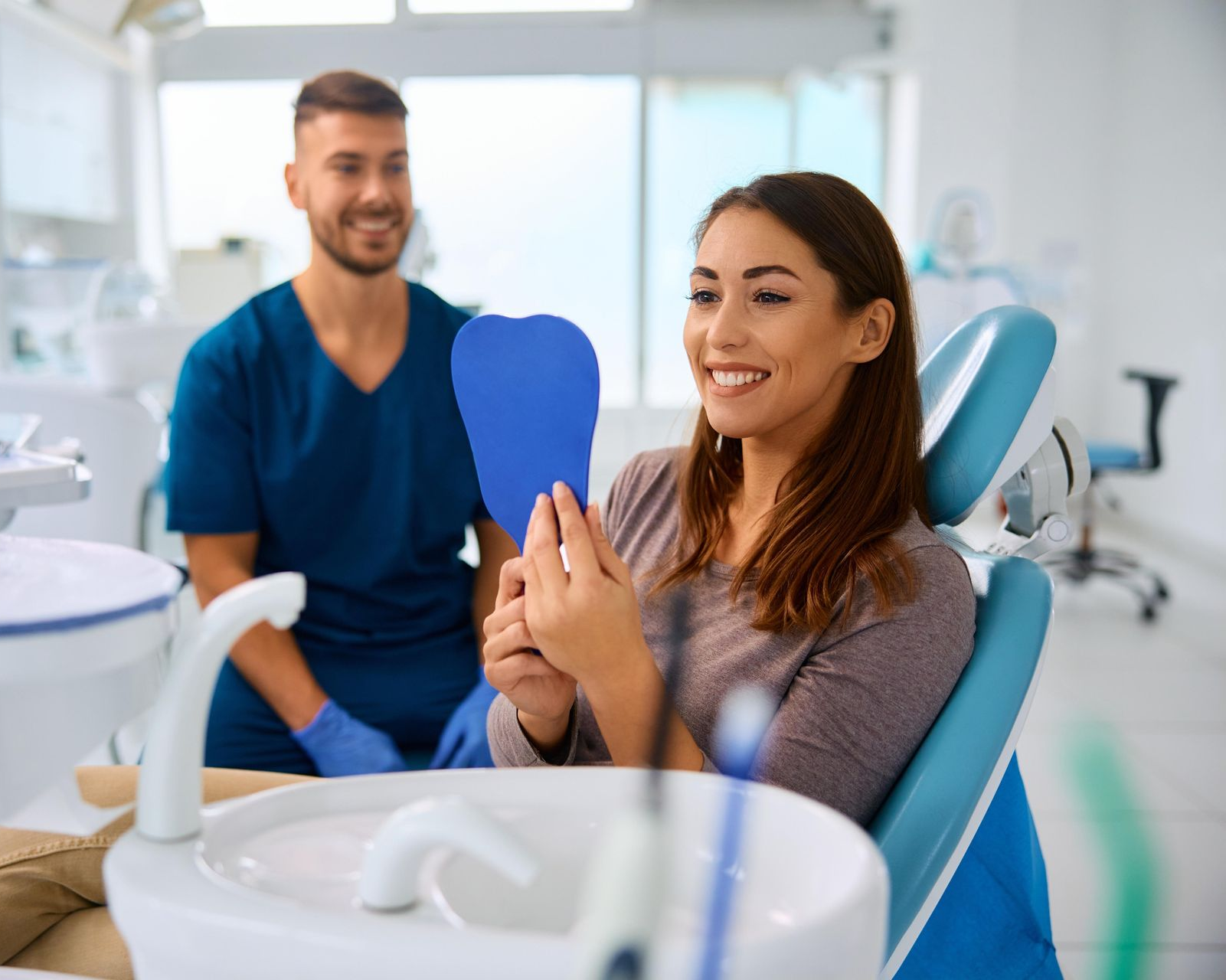 A person sitting in a dental chair smiles while looking into a handheld blue dental mirror with a clinician behind them.
