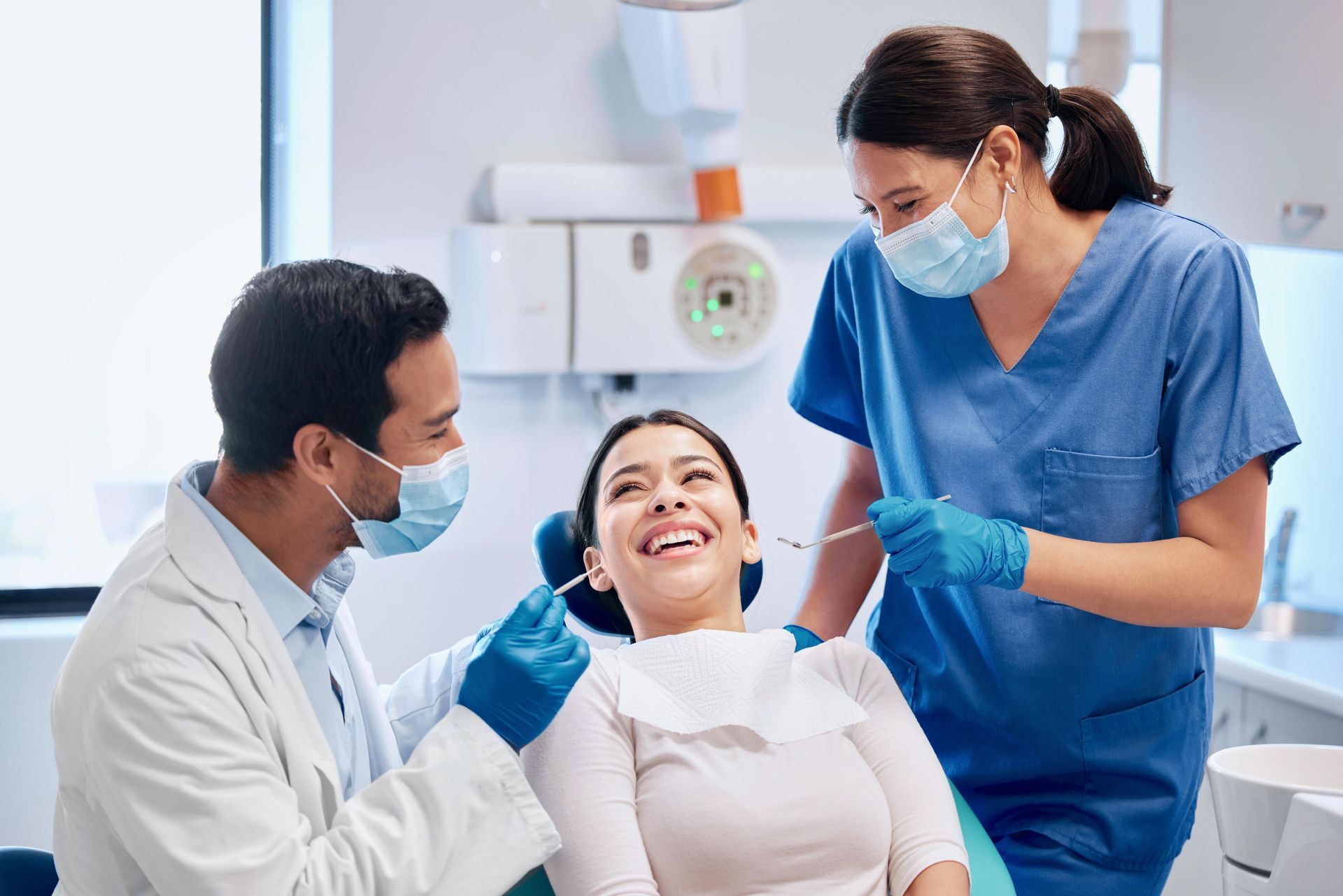 A dentist and assistant in medical scrubs and masks examine a smiling patient in a dental chair.