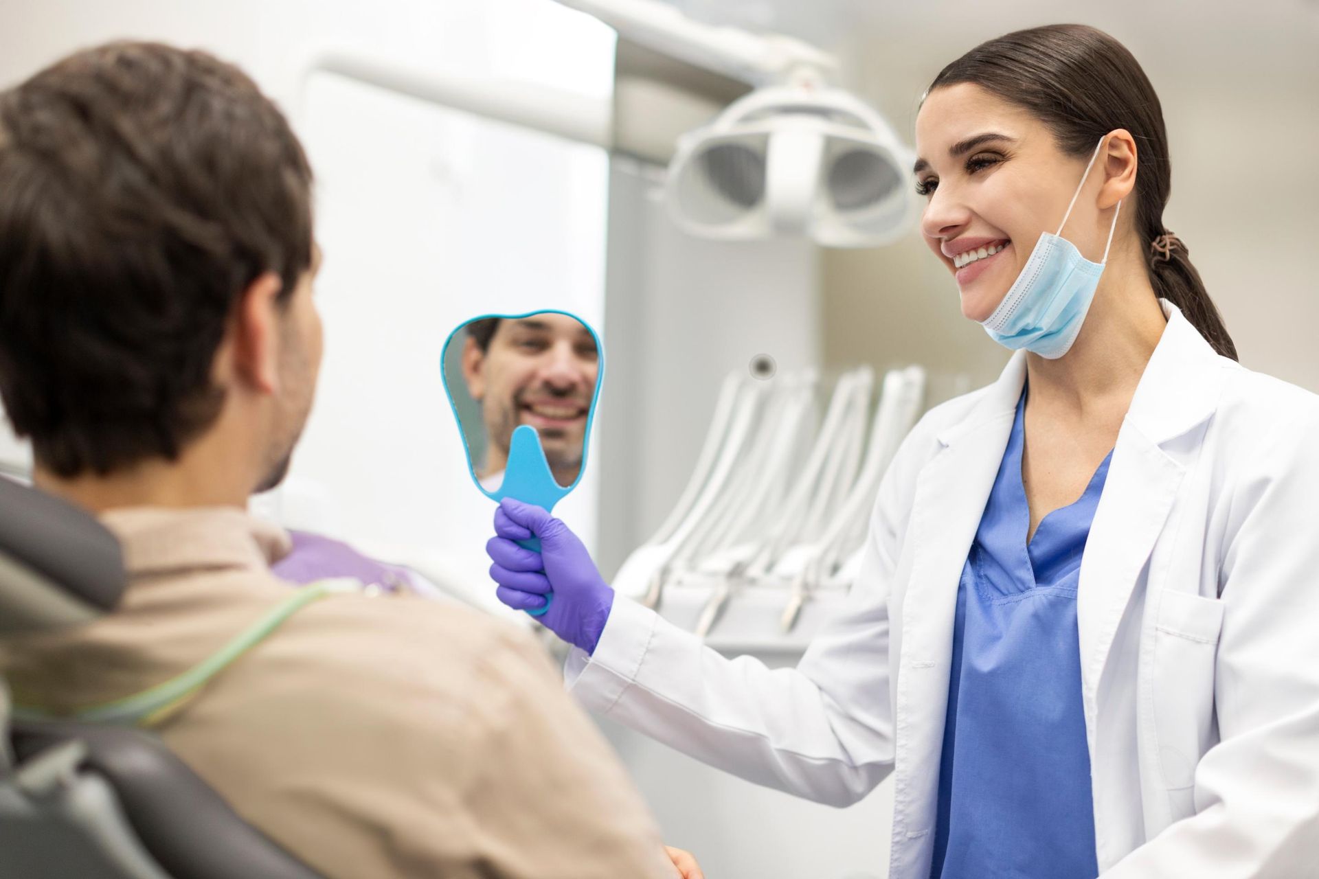 A dentist in a white coat and blue scrubs holds a hand mirror for a patient to see their smile in a dental office.