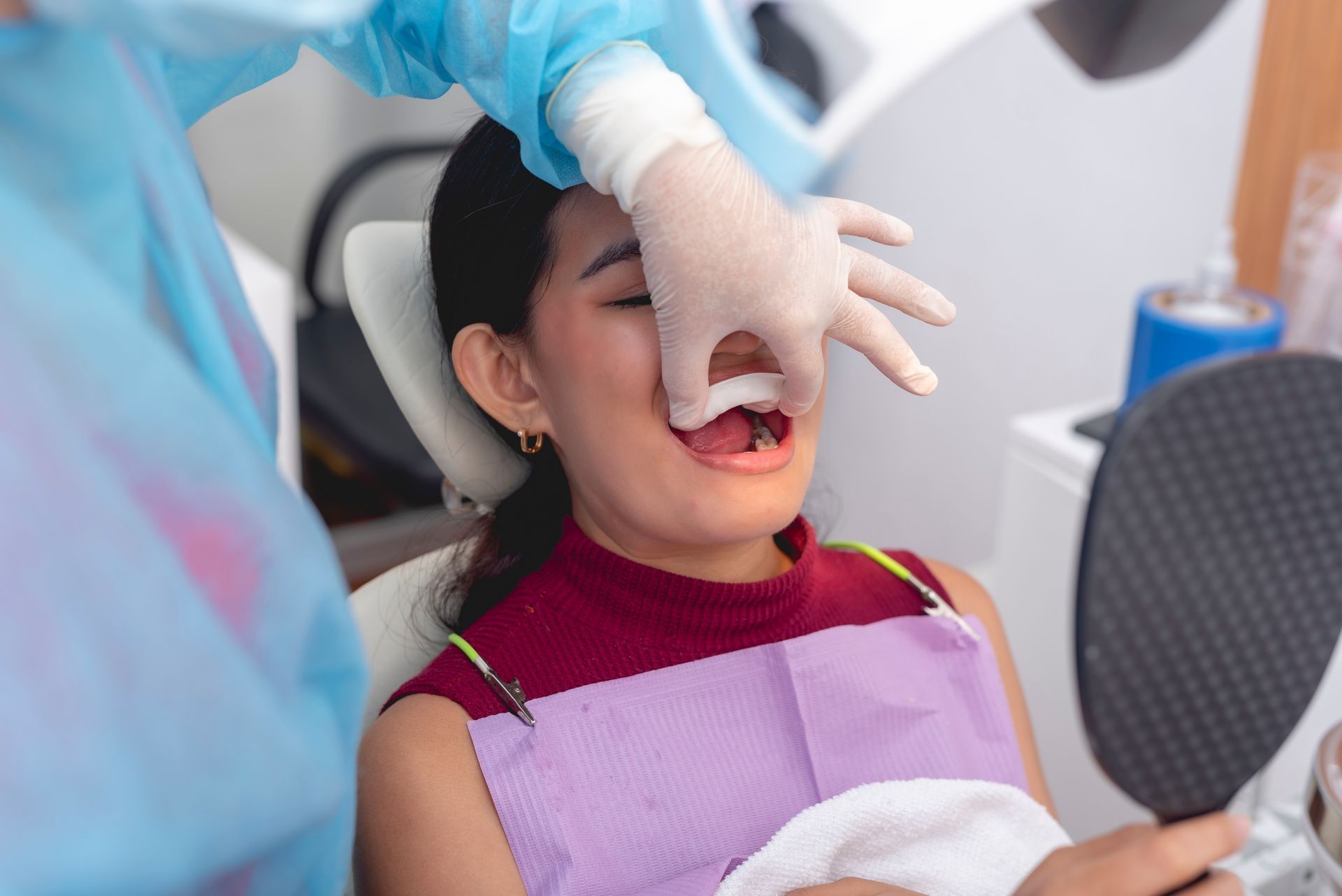 A dental professional wearing gloves adjusts a patient’s lip while they look into a hand mirror in a clinic.