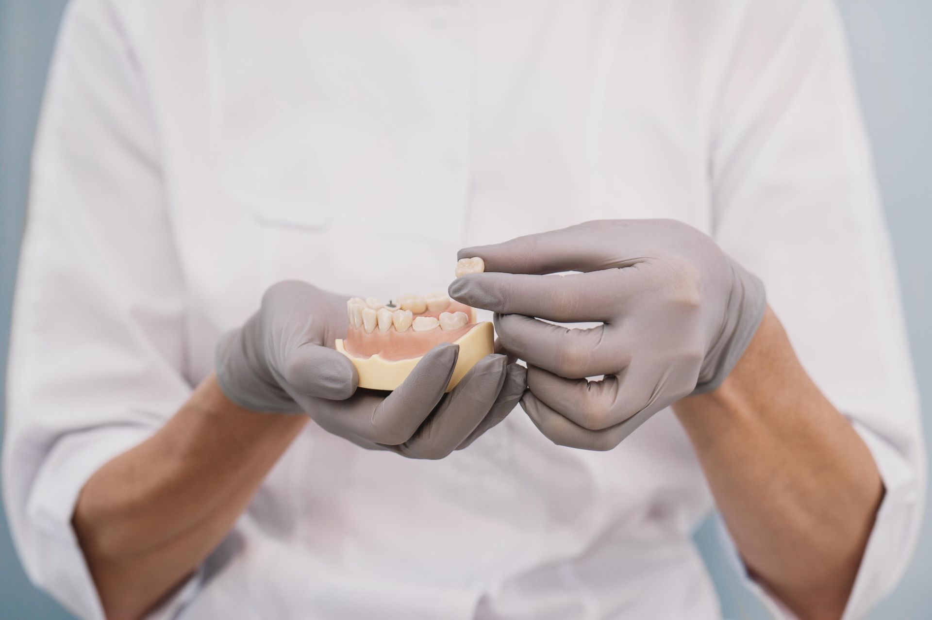 A dental professional in a white coat and gray gloves holds a dental model and a prosthetic tooth.