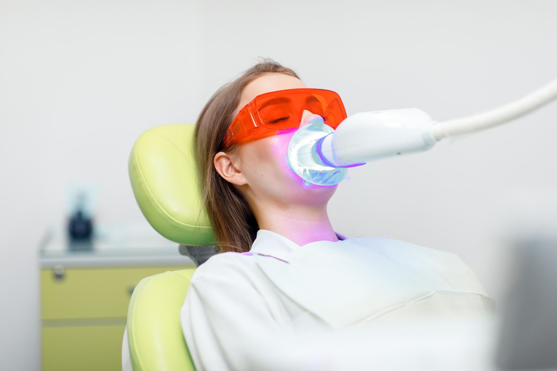 A person sits in a dental chair, wearing orange protective glasses, while receiving a professional teeth whitening treatment.