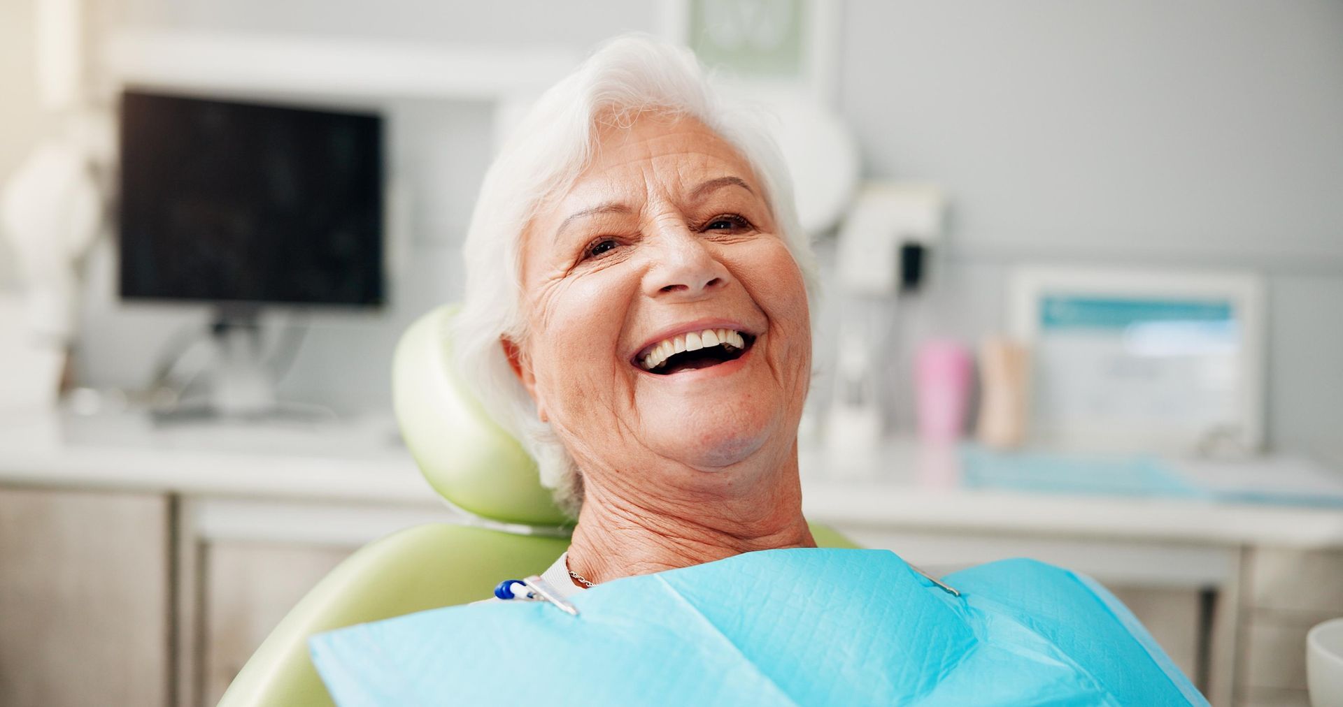 Smiling person in a dental chair wearing a blue bib, with blurred office equipment in the background.