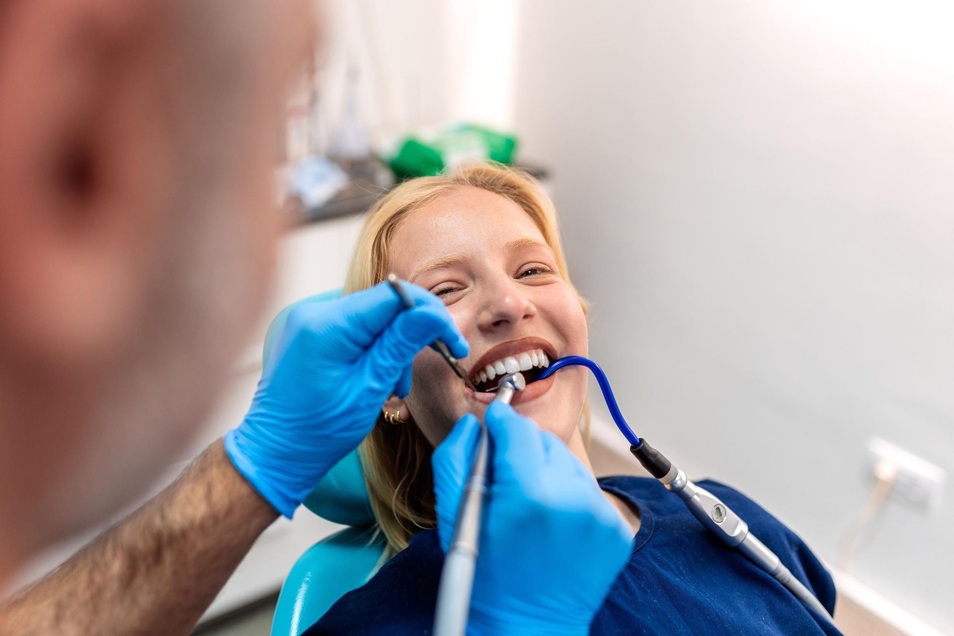 A dentist in blue gloves uses dental tools to examine a smiling patient in a clinical office setting.