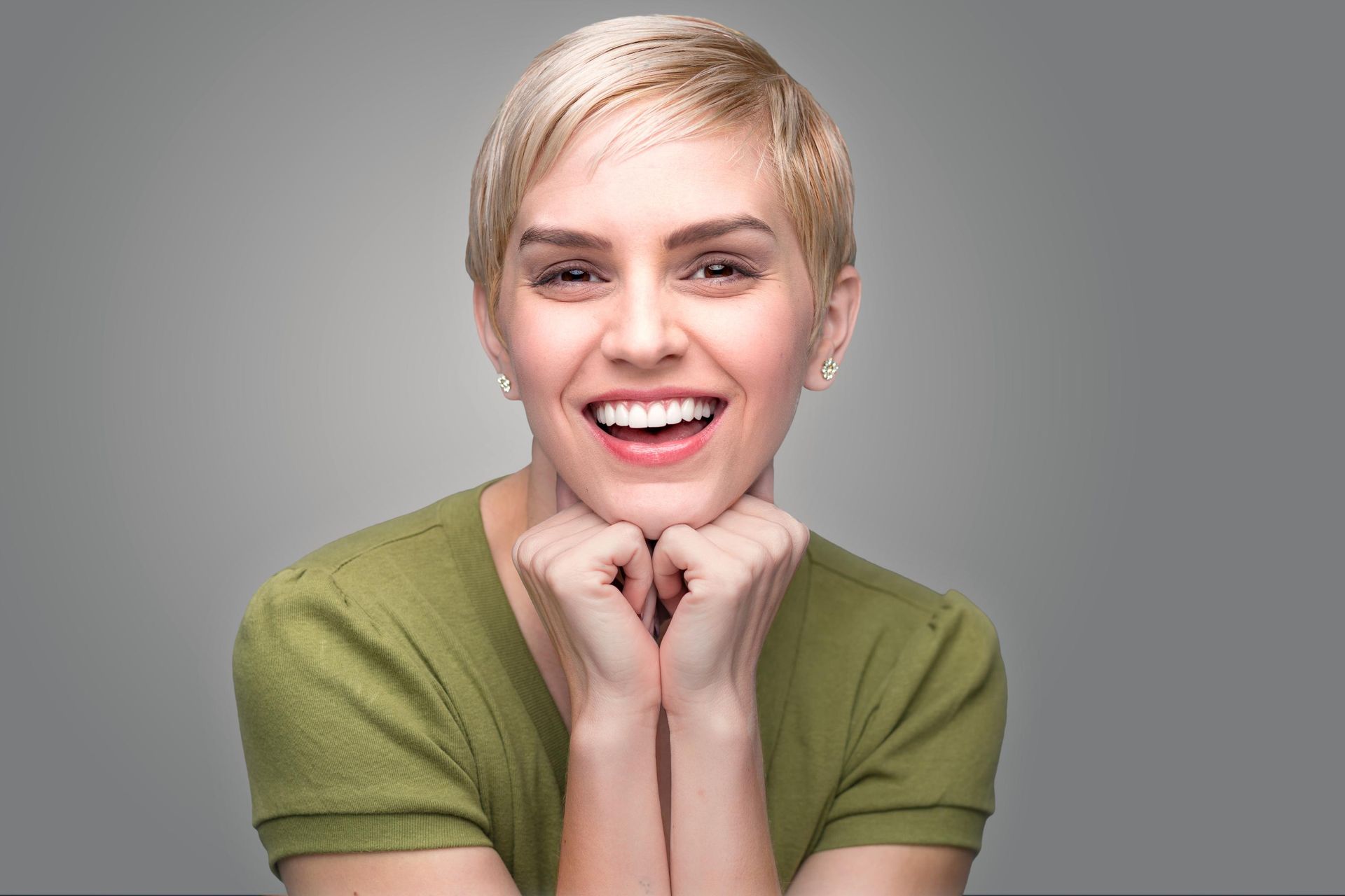 A person with a short blonde haircut and a green shirt smiles brightly, resting their chin on their folded hands.