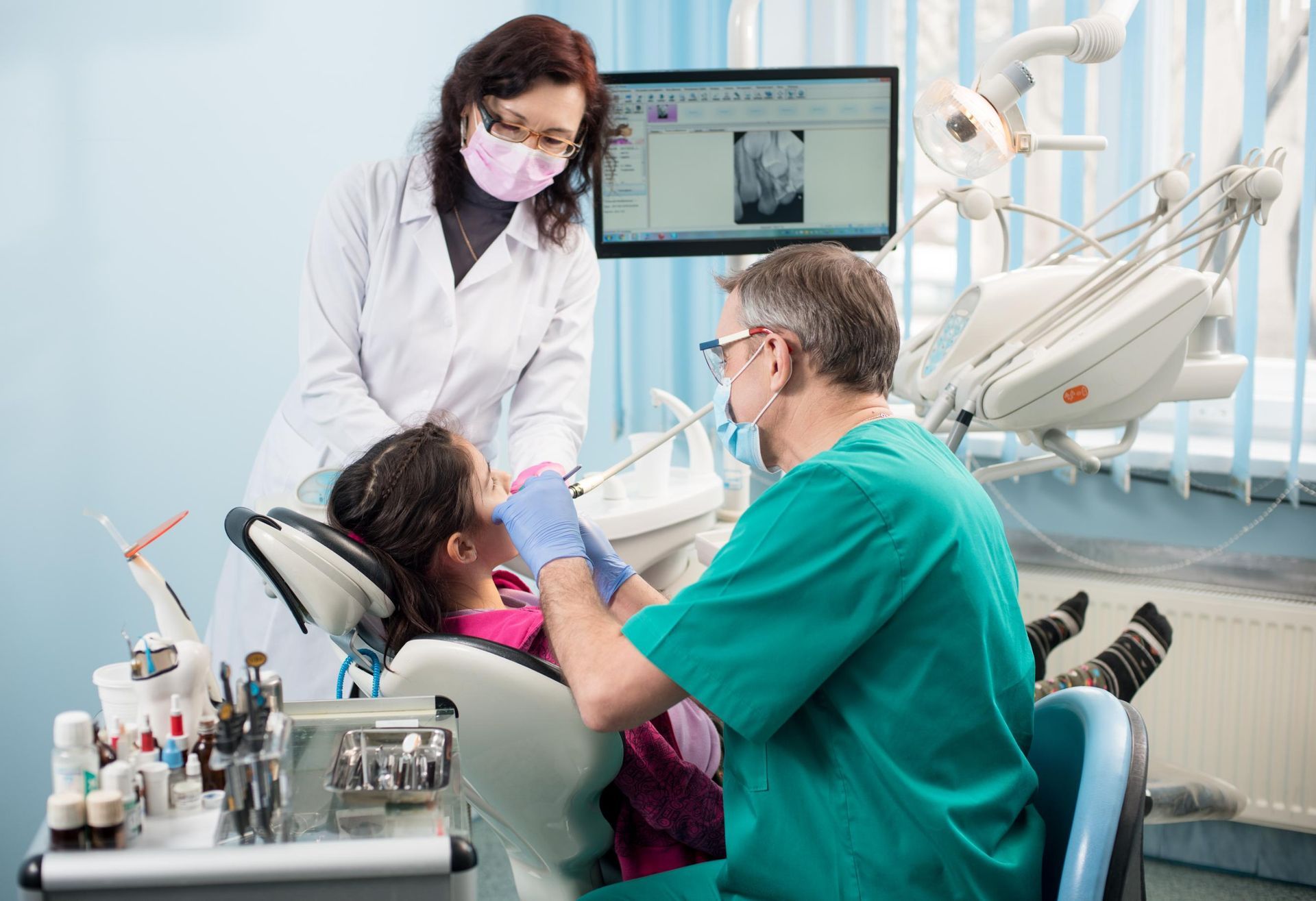 A dentist and an assistant perform a procedure on a patient in a dental chair, with a dental X-ray displayed on a monitor.