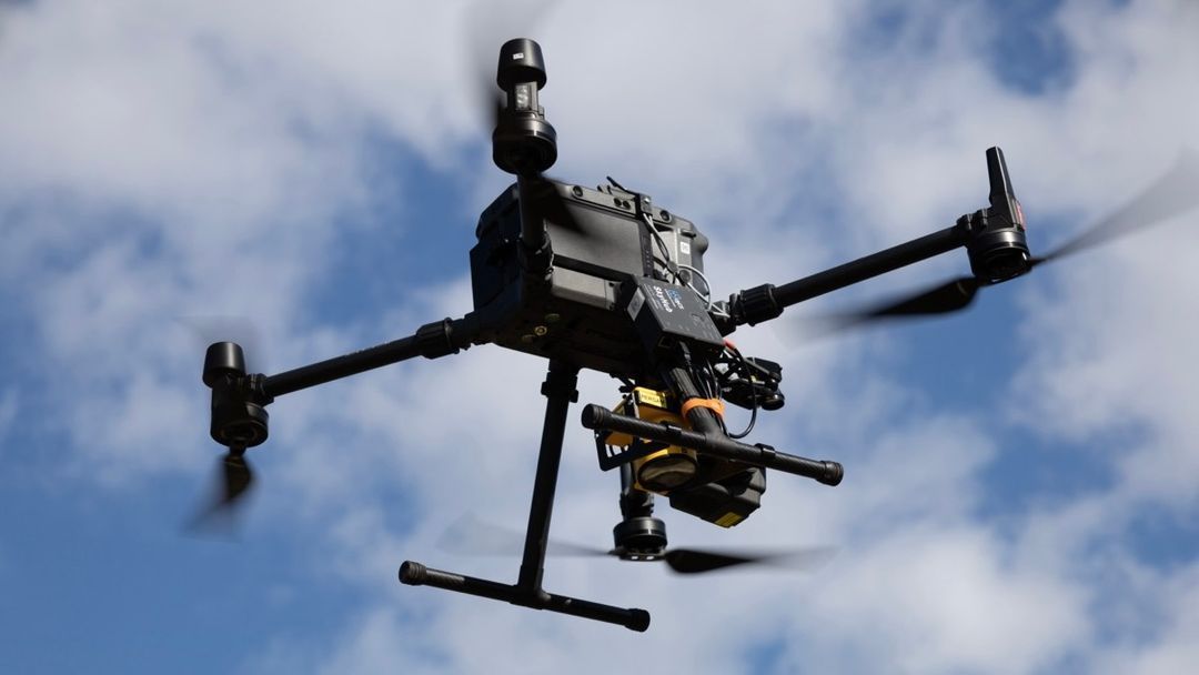 A drone is flying in the air with trees in the background.