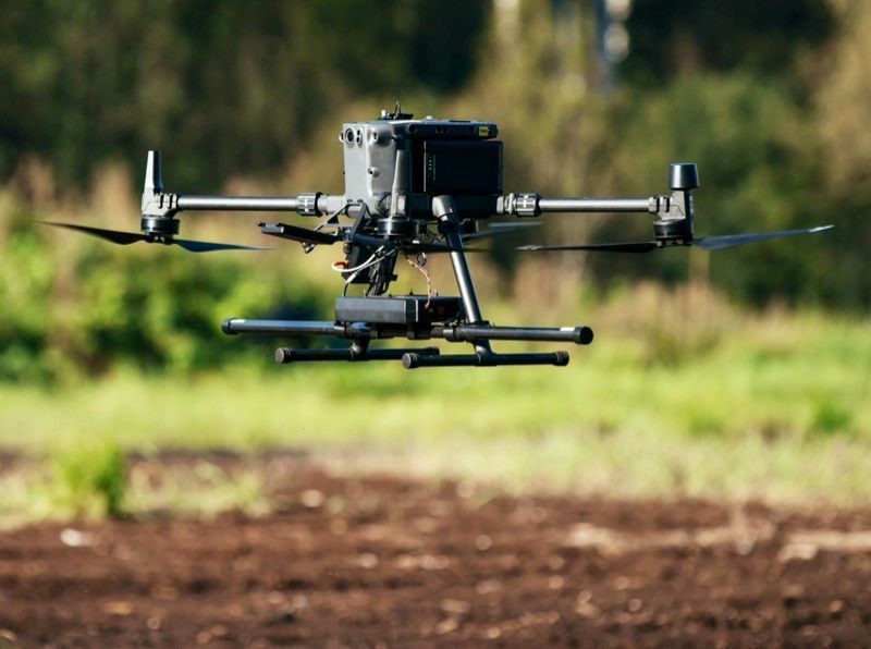 A drone is flying over a field with trees in the background.