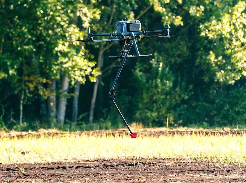 A drone is flying over a field with trees in the background.