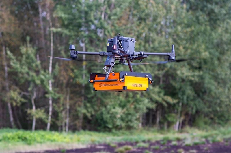 A drone is flying over a field with trees in the background.
